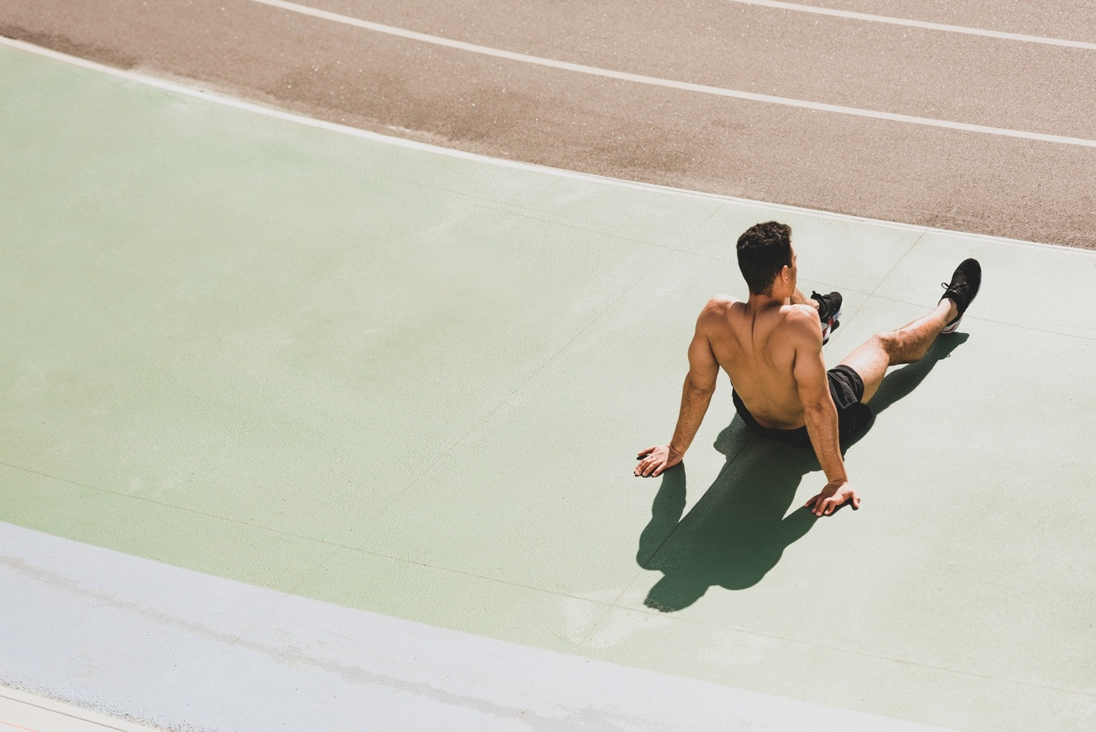 A man sitting on a sunlit outdoor court, stretching his legs and taking a quiet break after physical activity.