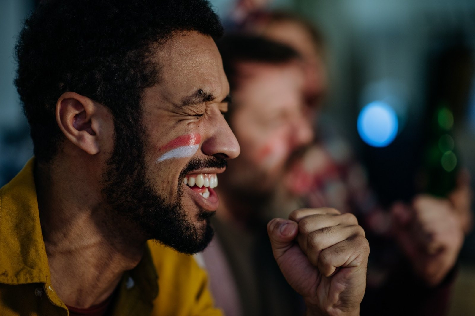 Man cheering with his eyes closed and fist raised, with another person blurred in the background.