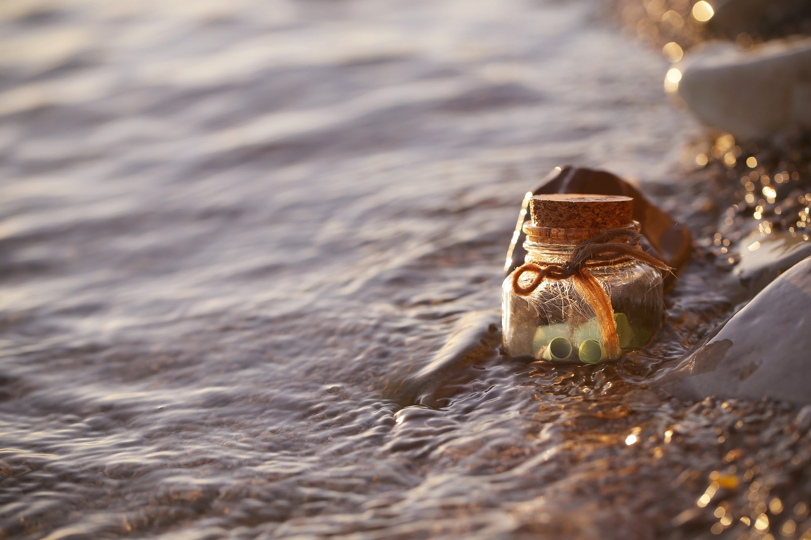 A small glass bottle with a cork lid resting at the edge of gently flowing water, surrounded by smooth stones and warm sunlight.