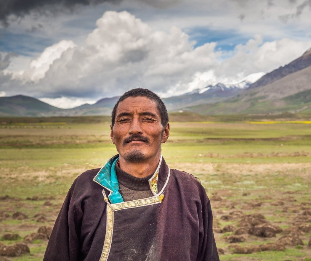 Man wearing traditional clothing standing in an open grassland with mountains and cloudy skies behind him.