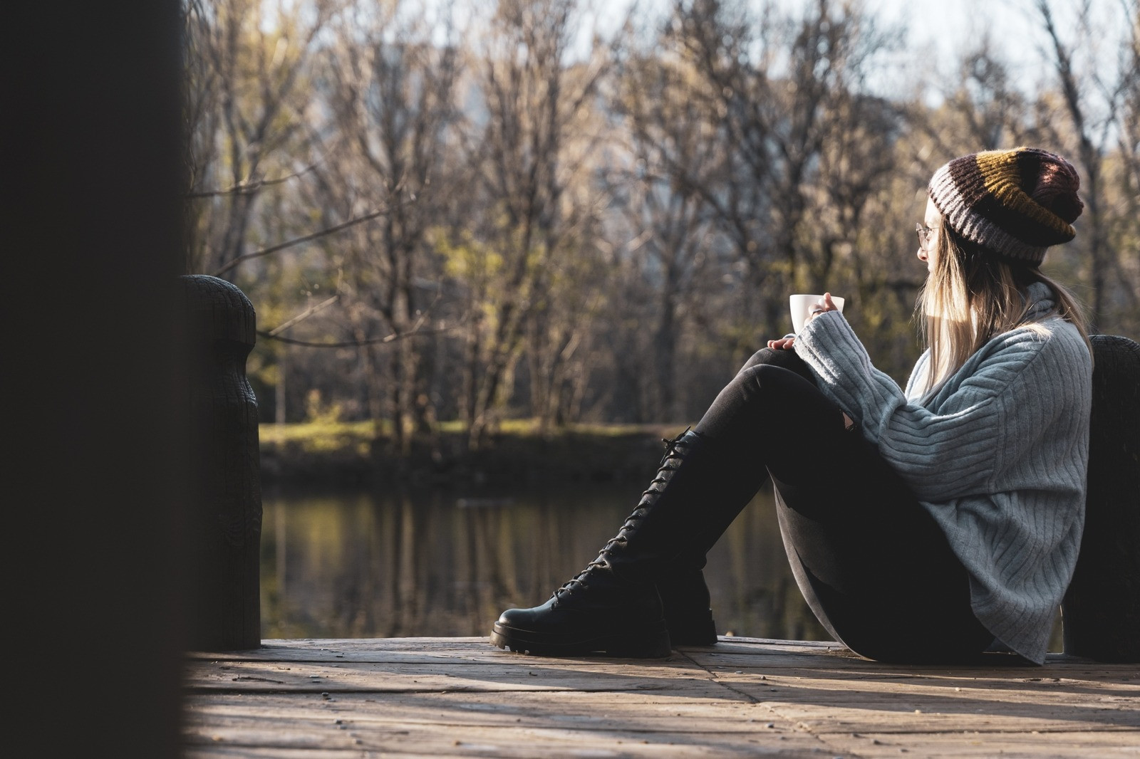 Woman sitting by a lake with a warm drink, quietly reflecting in a peaceful outdoor setting.