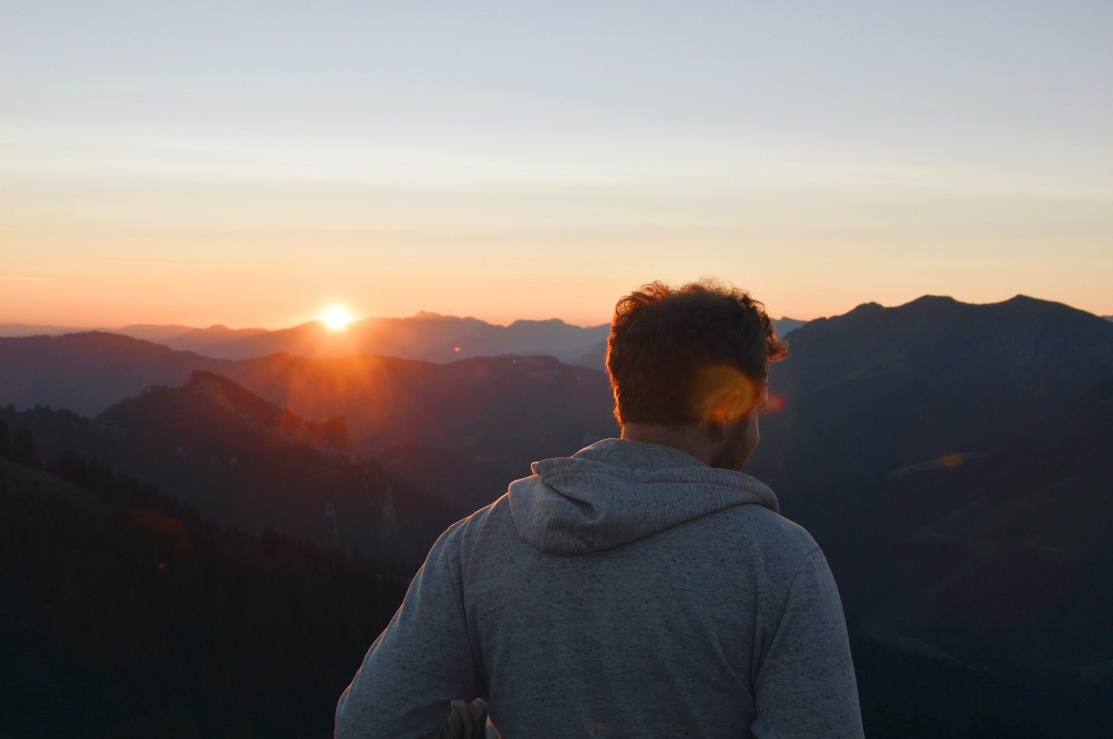 Man wearing a gray hoodie looking toward a sunset over a mountain landscape.