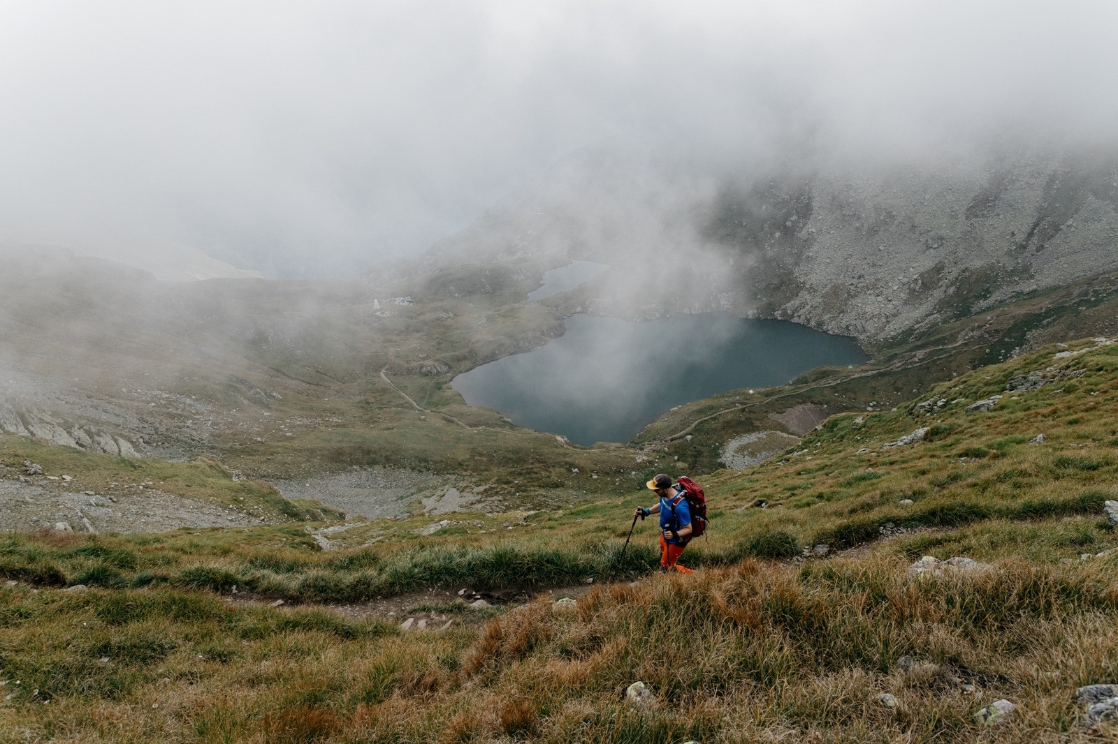 A hiker carrying a backpack walks along a grassy mountain trail overlooking a mist-covered lake, symbolizing gradual change and emotional processing.