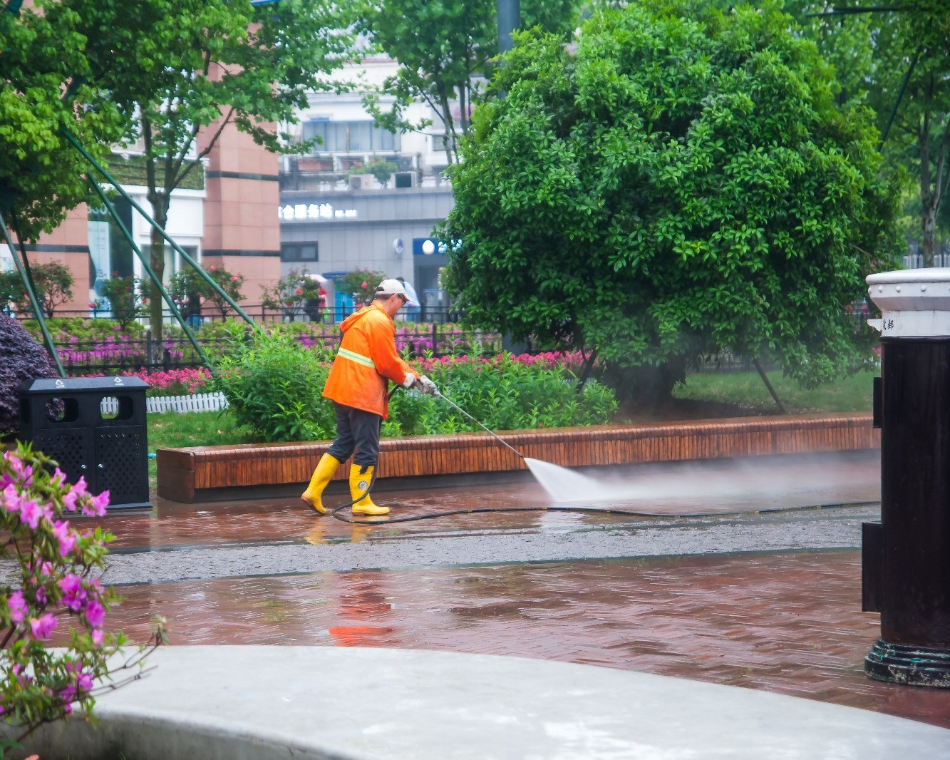 Park maintenance worker in an orange safety jacket and yellow boots pressure-washing a paved walkway surrounded by green trees and flowers.