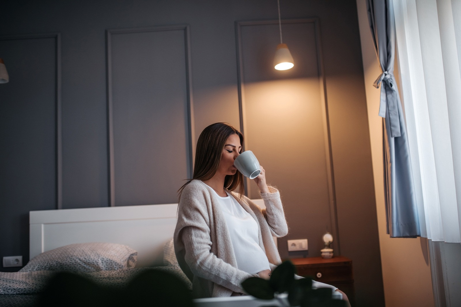 Pregnant woman sitting on a bed and drinking from a cup in a softly lit bedroom