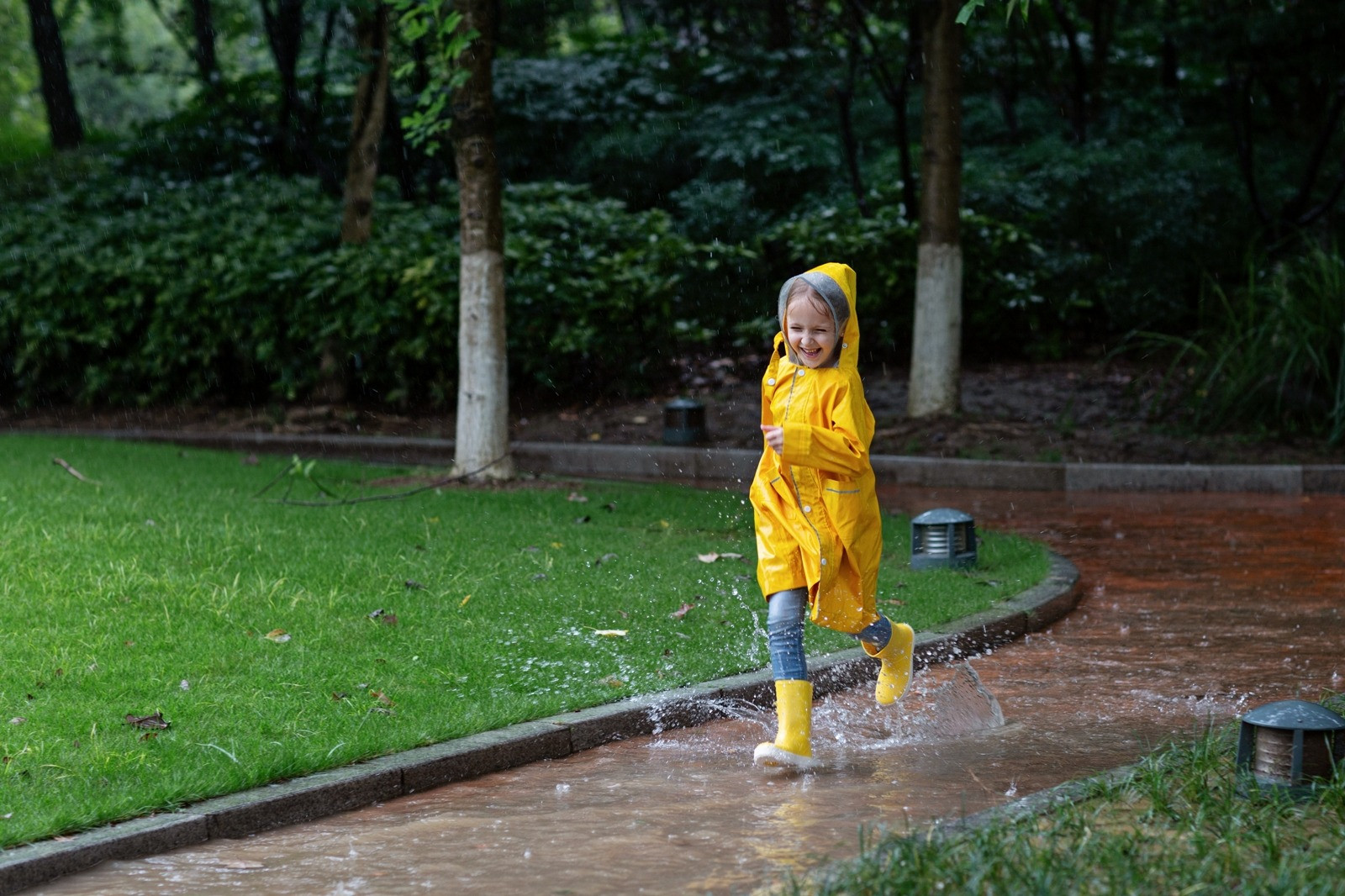 A smiling child wearing a yellow raincoat and boots splashes through puddles on a park pathway, illustrating movement and changing physical sensations.