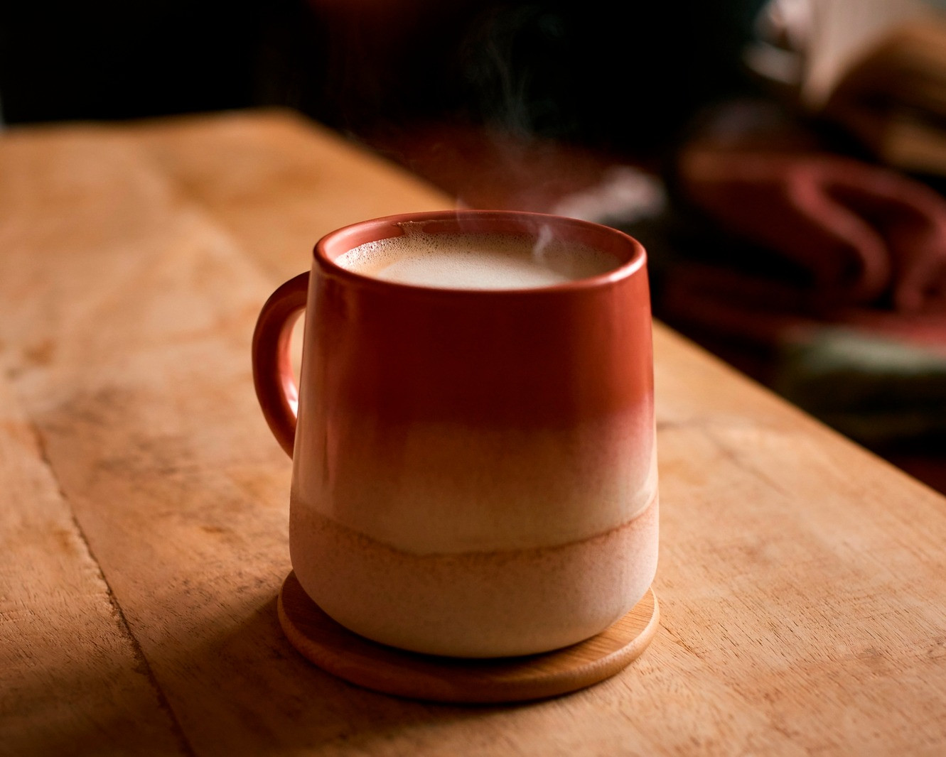 Steam rising from a red and cream ceramic mug on a wooden coaster and table.
