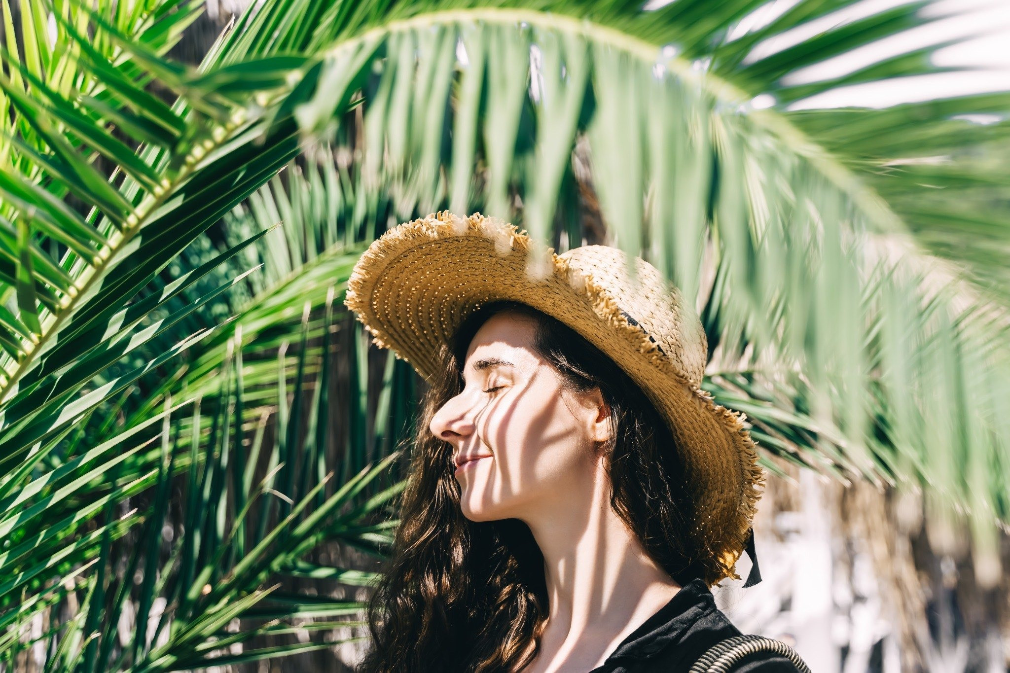 Woman wearing a straw hat standing among palm leaves with her eyes closed
