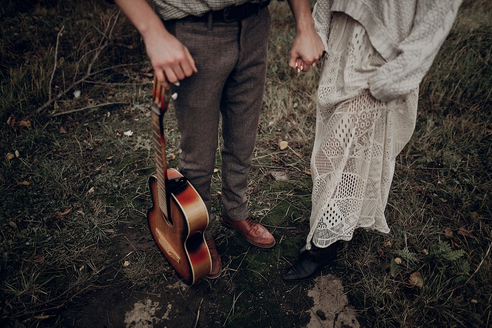 Two people holding hands outdoors, one carrying an acoustic guitar and the other wearing a cream lace dress.