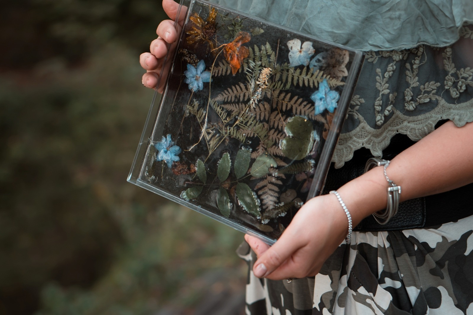 Close-up of a person holding a glass frame containing preserved flowers, fern leaves, and natural botanical arrangements.