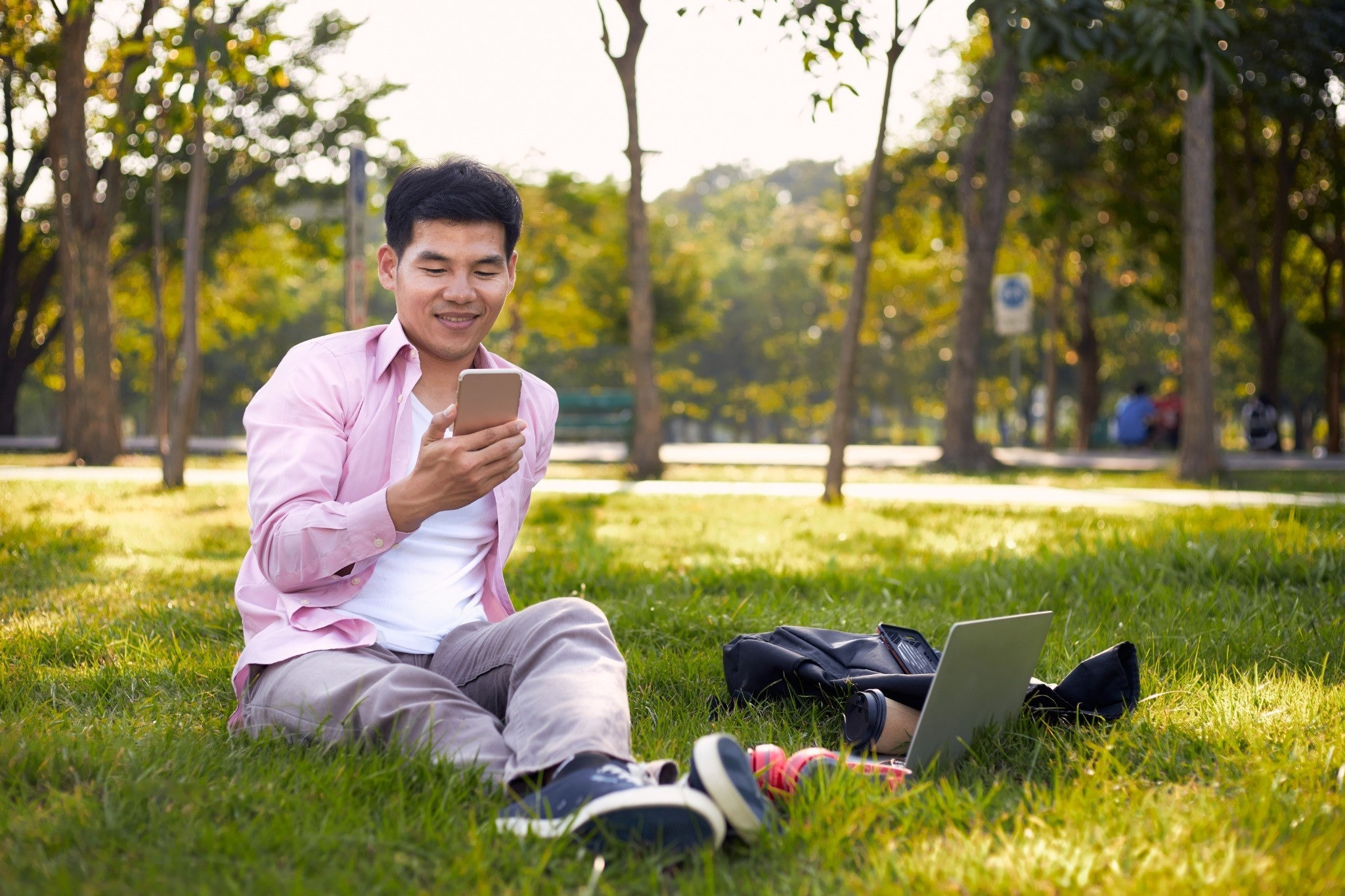 Man sitting on the grass in a park using his phone beside a laptop