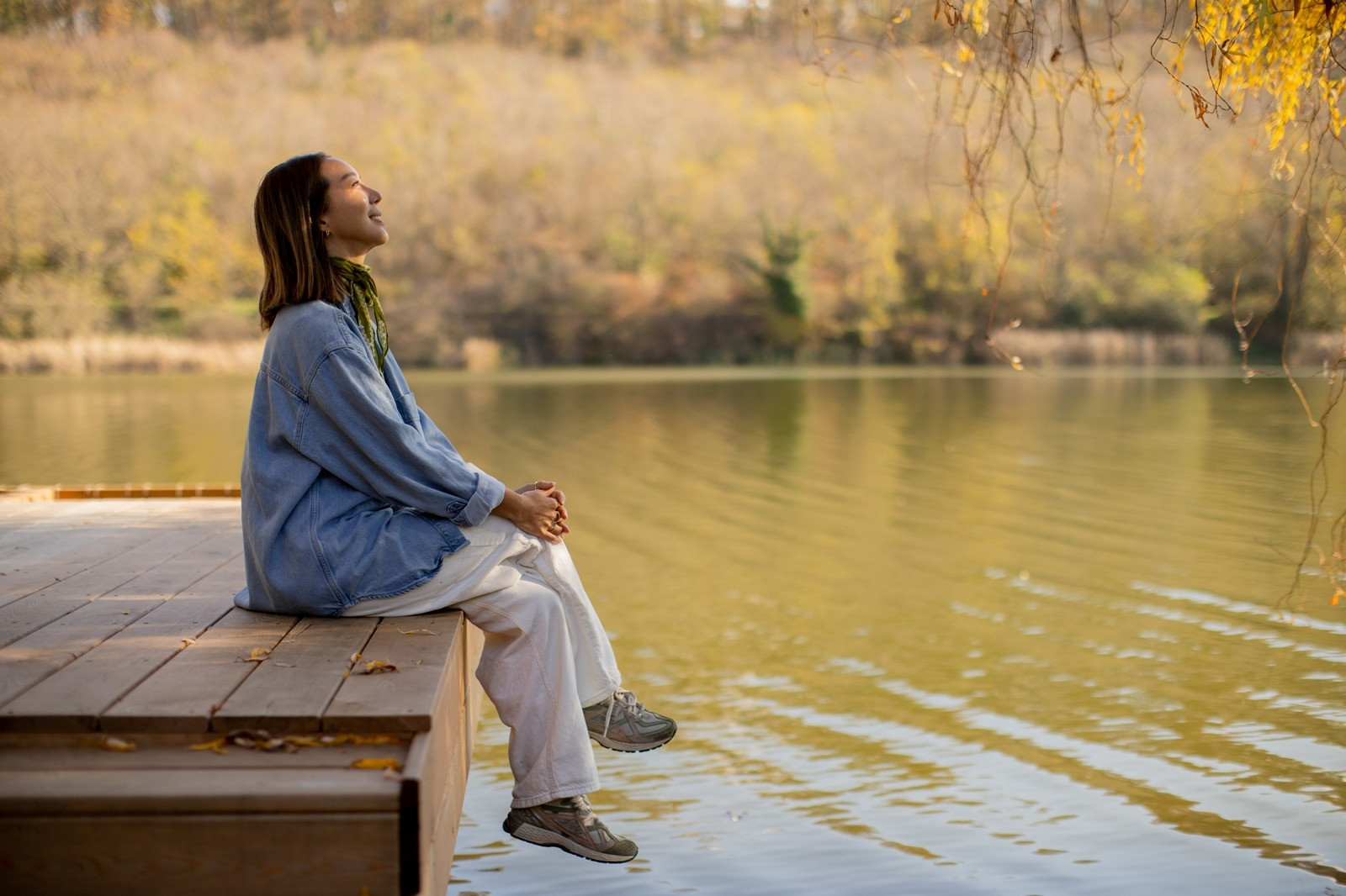 Woman sitting calmly beside a lake in a peaceful autumn setting, reflecting outdoors.