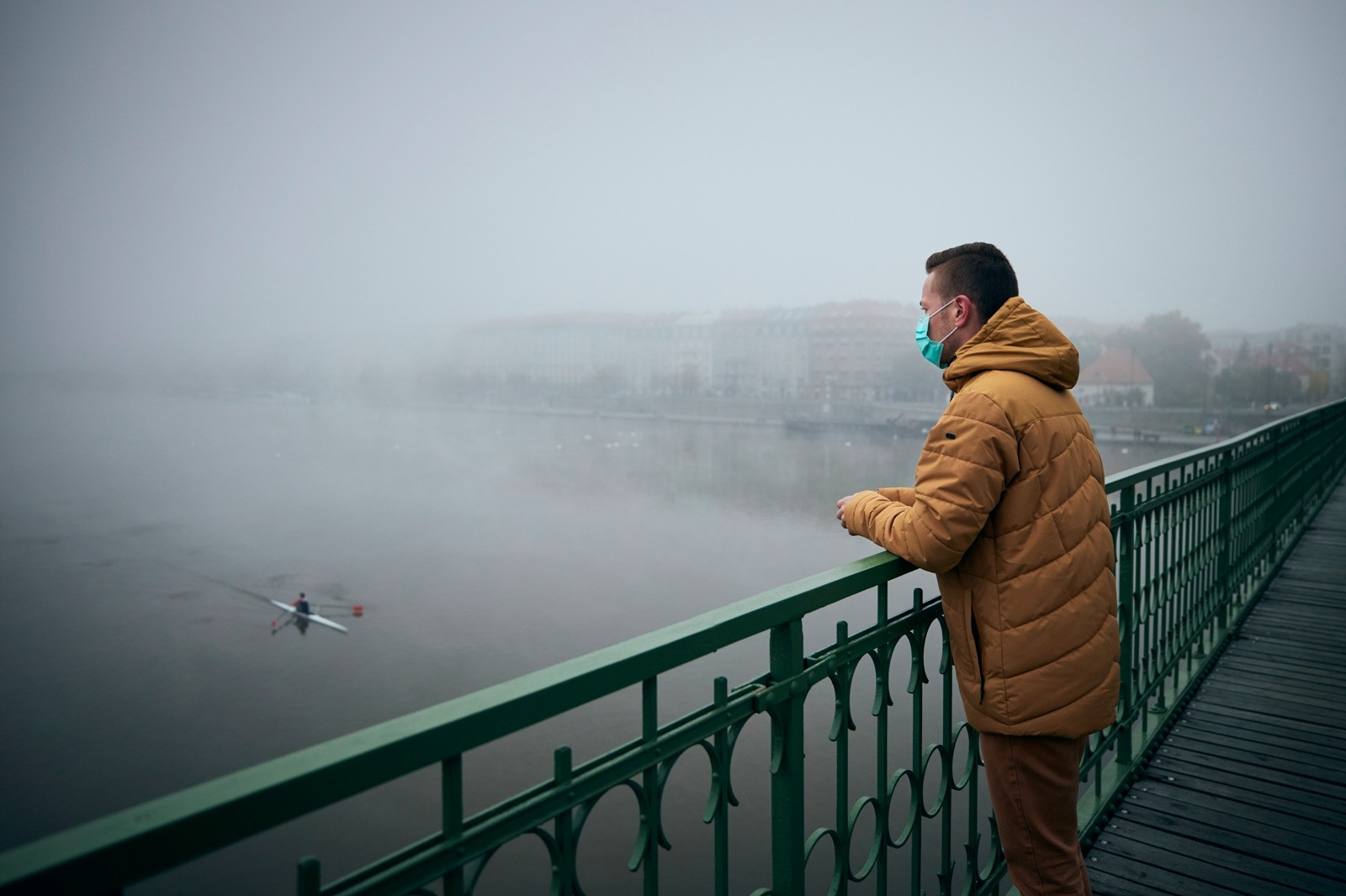 A man wearing a protective face mask and brown jacket stands on a bridge overlooking a foggy river, reflecting on his surroundings.