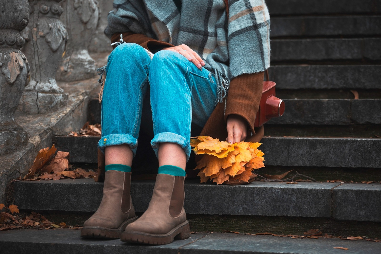Person sitting on outdoor stone steps wearing a blue-gray scarf and jeans, holding a bunch of yellow autumn leaves in a quiet, reflective moment.