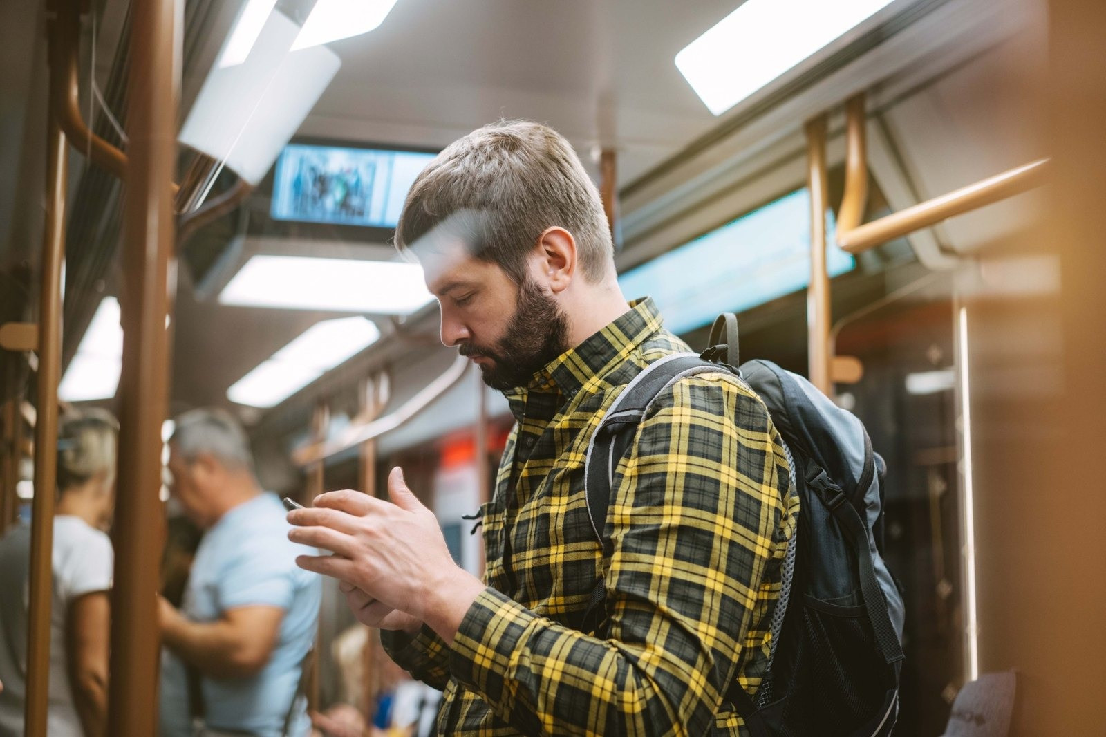 Bearded man wearing a yellow plaid shirt and backpack looking down at his smartphone while riding public transportation.