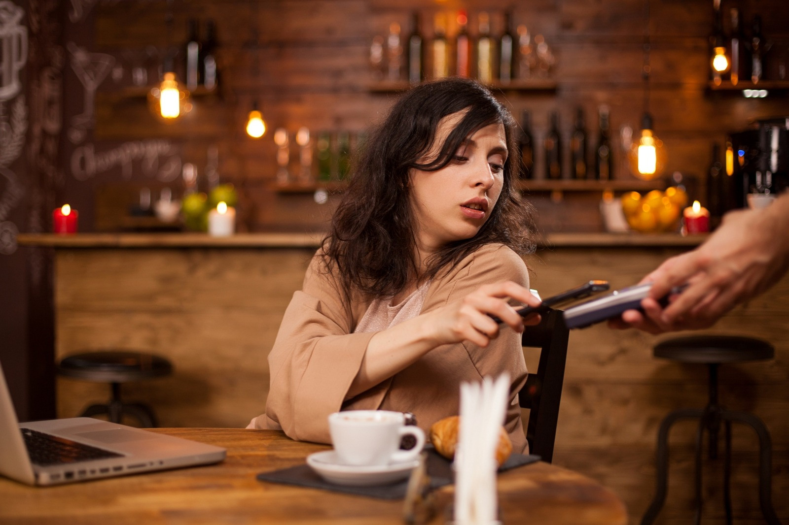 Woman seated at a café table using a payment terminal beside a laptop and cup of coffee.