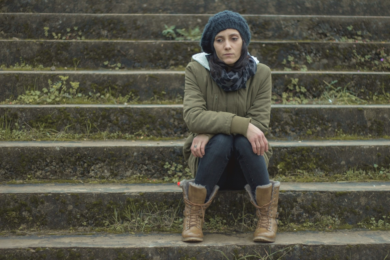 Woman in winter clothing sitting thoughtfully on outdoor stone steps, wearing a knit hat, green jacket, and brown boots.