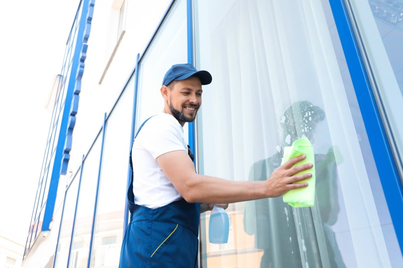 Smiling window cleaner wearing a blue cap and work overalls, wiping a large glass window with a green cloth.
