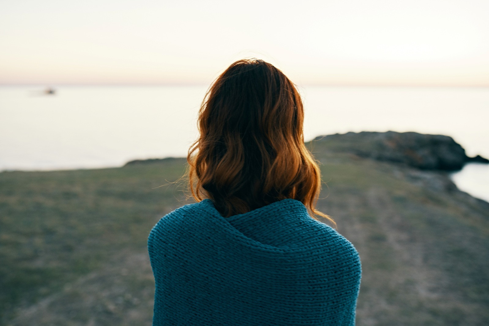 A person wrapped in a blue knitted blanket, viewed from behind, looking out over calm water from a rocky shore.