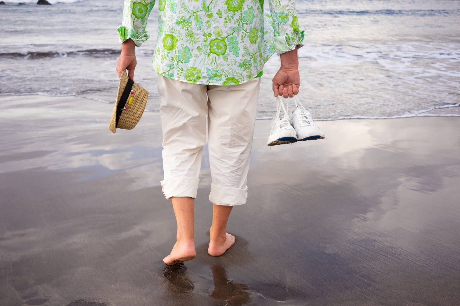 Person walking barefoot along the beach while carrying shoes and a sunhat.