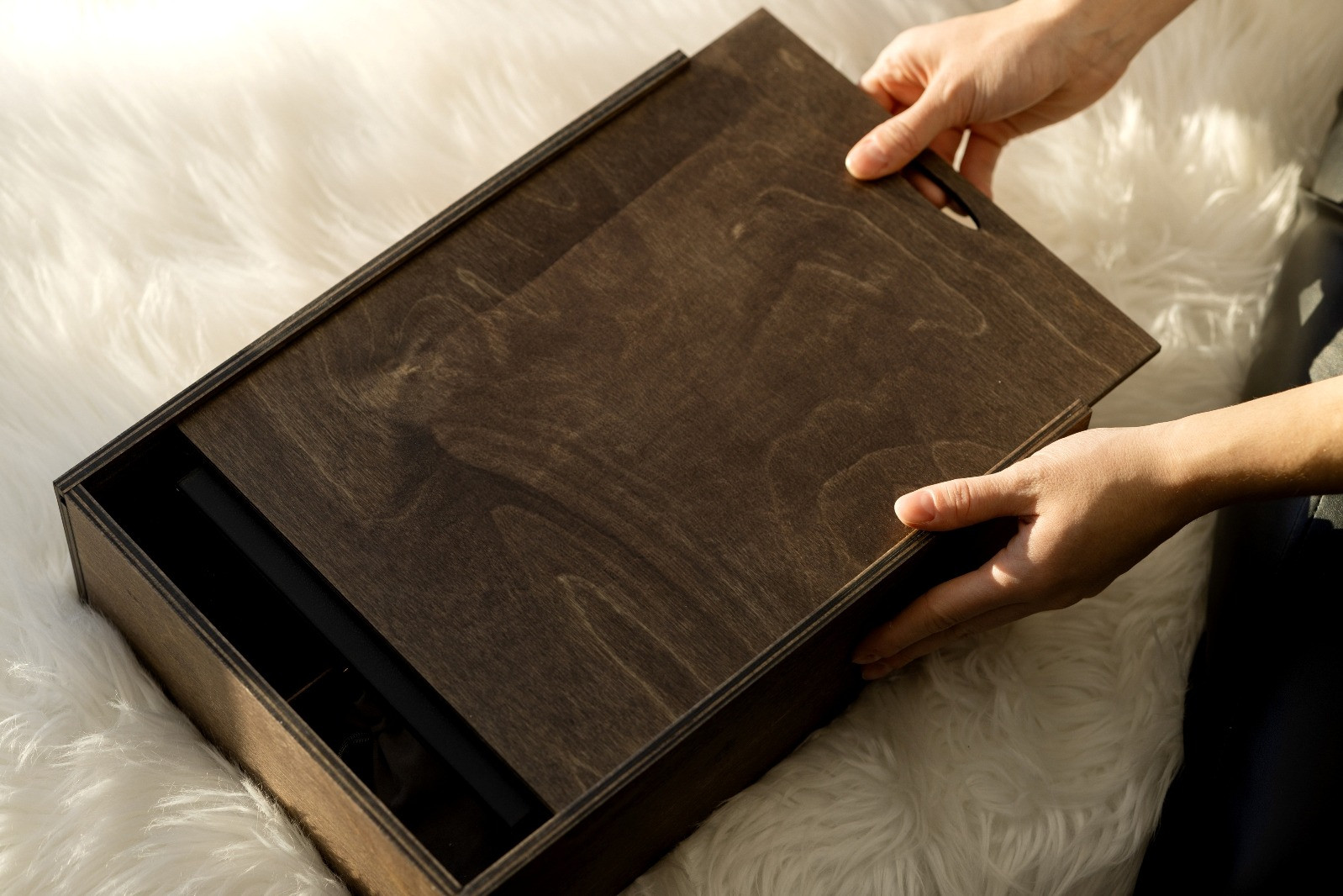 Hands lifting the lid of a dark wooden box resting on a white textured surface.