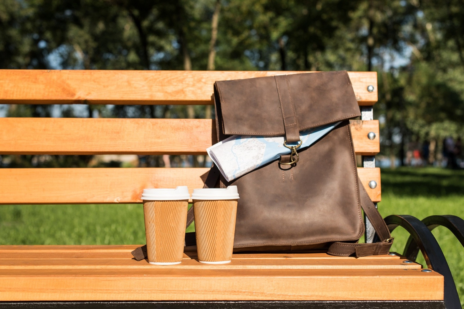 Two takeaway cups beside a brown leather bag with a folded map on a sunlit park bench.