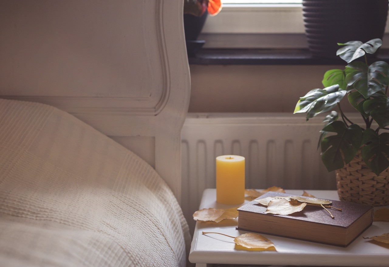 Cozy bedroom scene with a bedside table, candle, book, and indoor plants near a sunlit window.