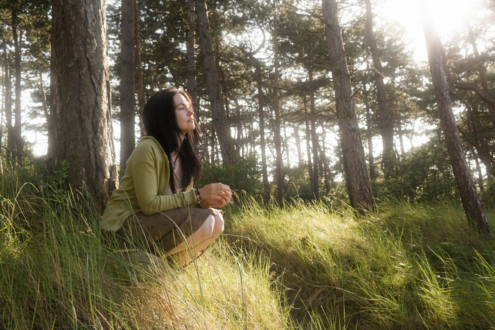 Woman sitting peacefully in a sunlit forest, with her eyes closed and hands gently resting together.
