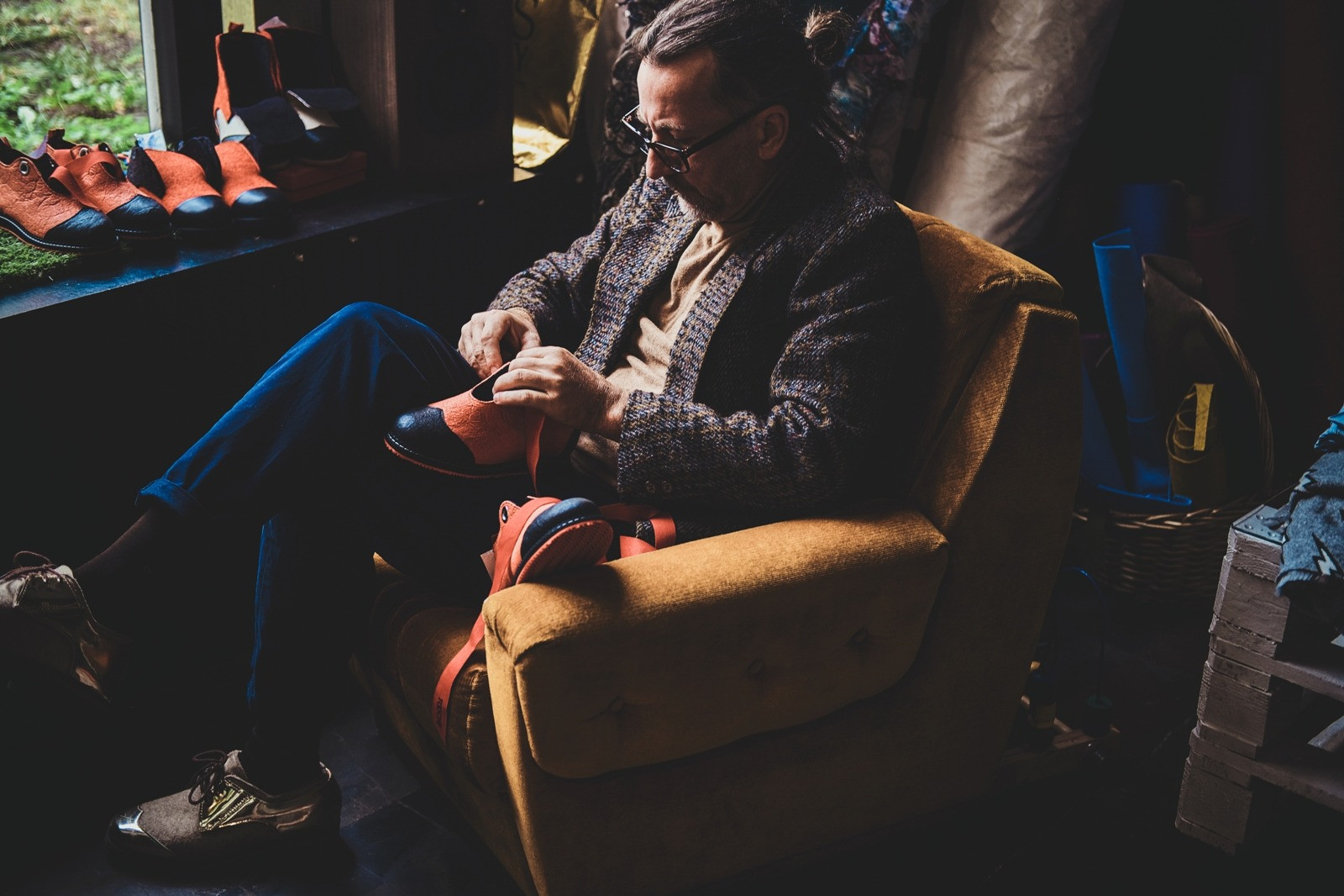 A man wearing glasses sits in a mustard-colored armchair by a window, working on an orange and black shoe.
