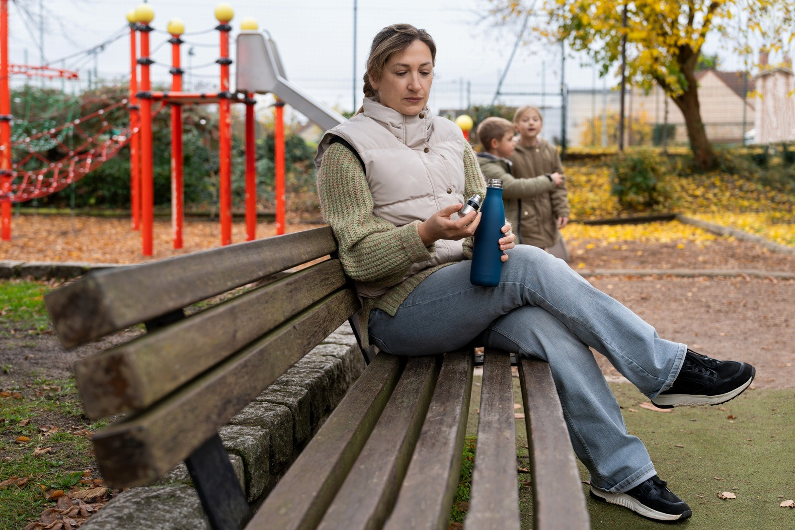 Woman sitting on a park bench near a playground, reflecting quietly while children stand in the background.
