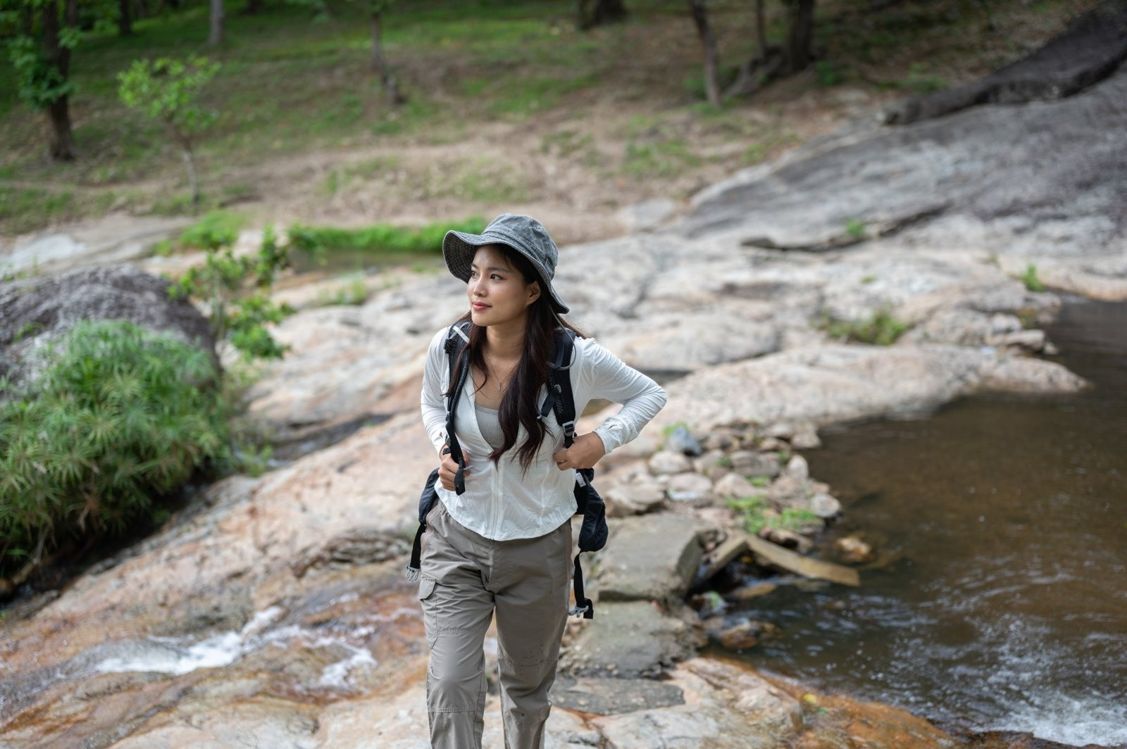 Woman wearing a gray bucket hat and backpack standing on rocks beside a stream in a wooded area.