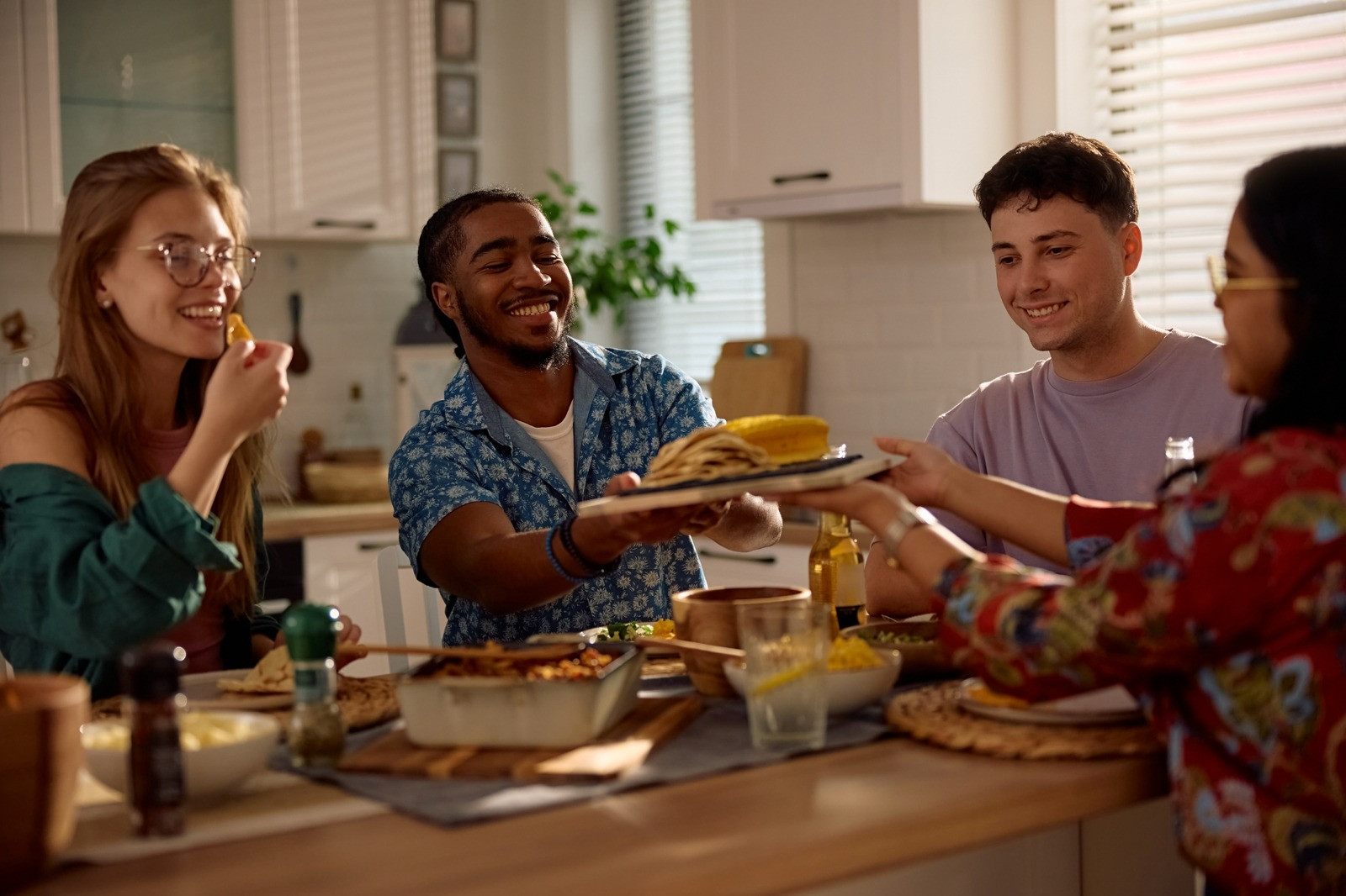 Four friends smiling and sharing food around a dining table in a bright kitchen, enjoying a relaxed social gathering.