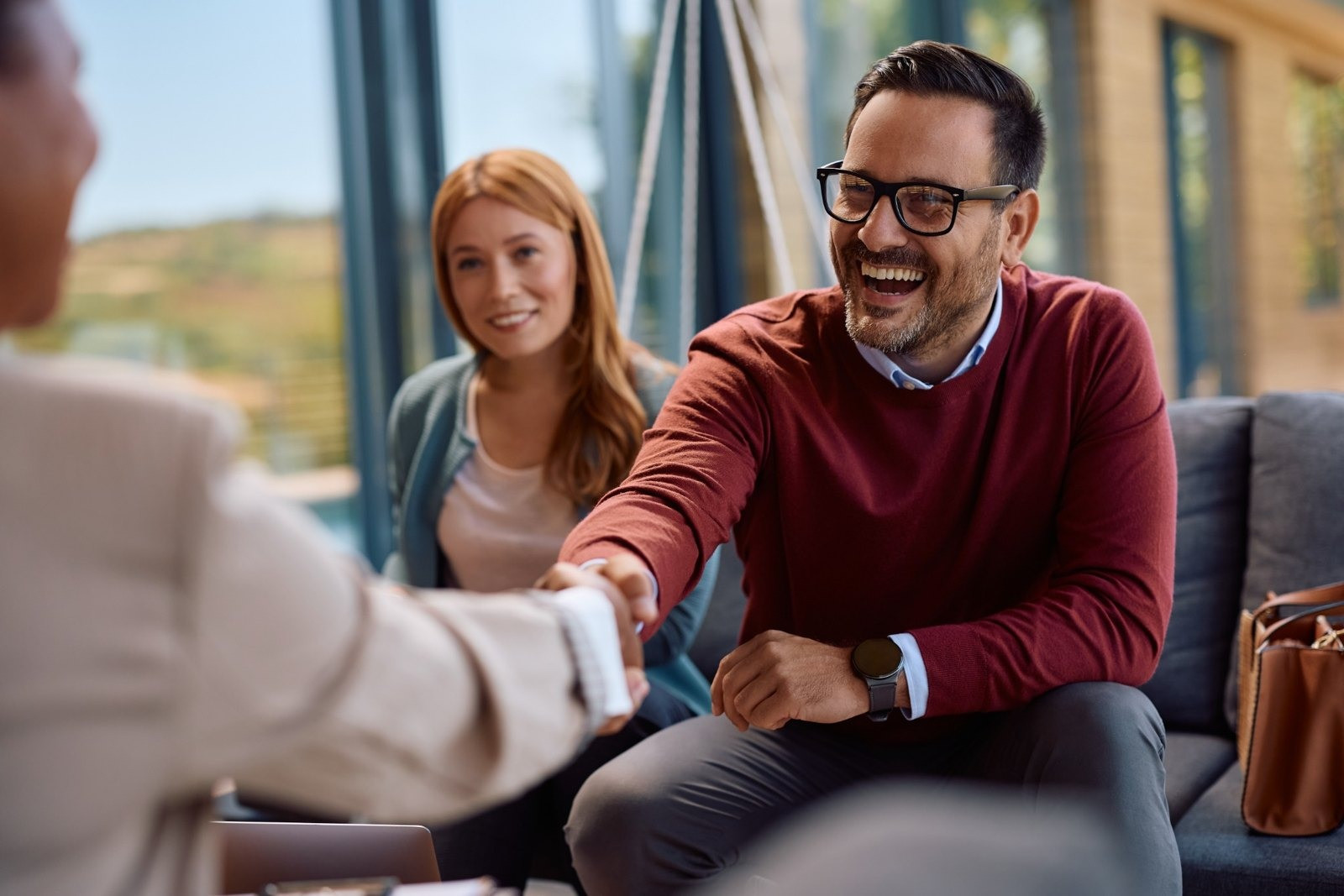 Smiling man wearing glasses and a burgundy sweater shaking hands with another person while a woman sits beside him.