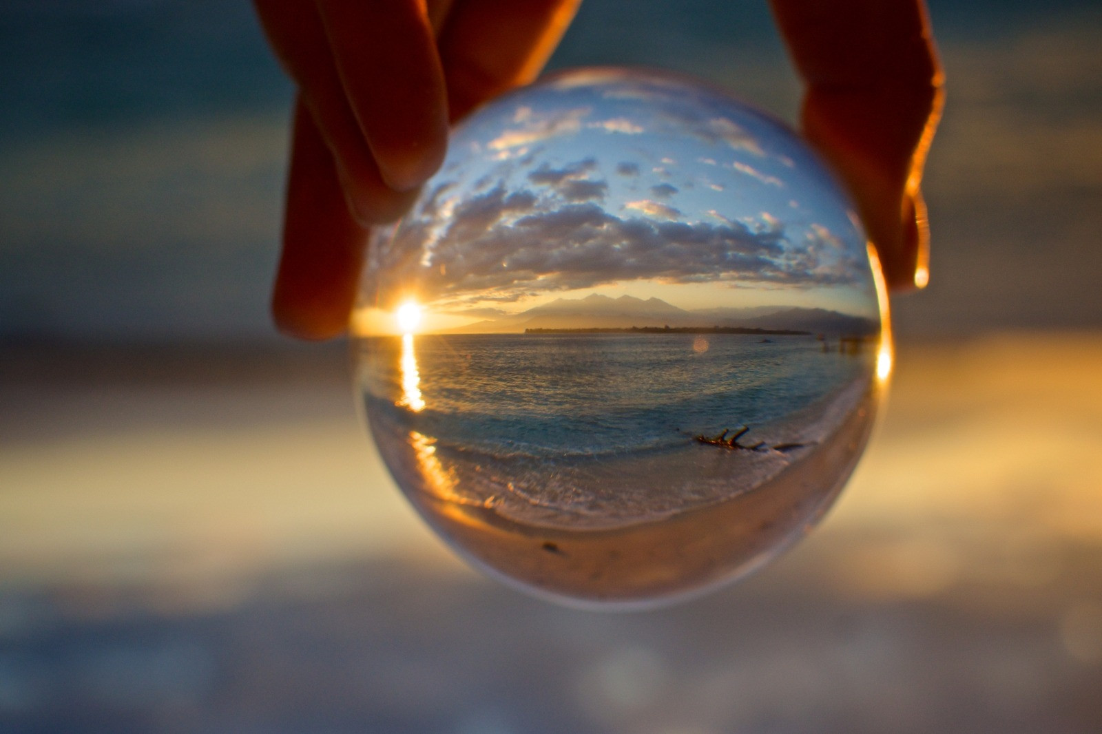 A hand holding a clear glass sphere showing a sunset over water, with distant mountains and clouds.