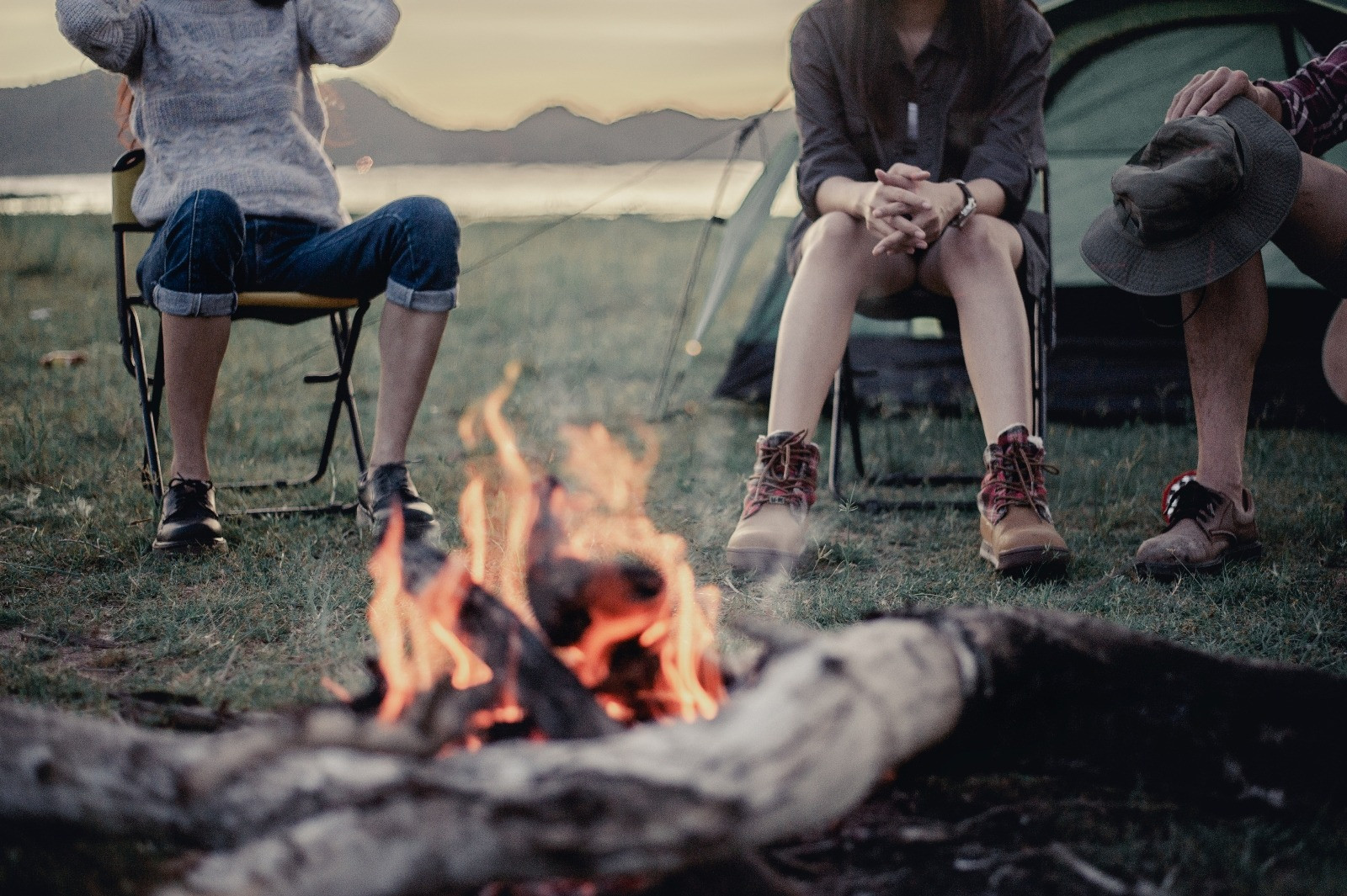 Three people sitting around a campfire at a campsite with tents and mountains in the background.
