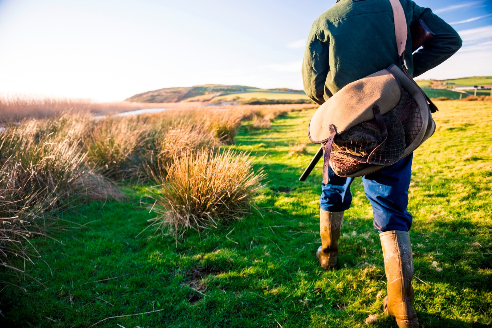 Person walking through a grassy countryside carrying a shoulder bag.