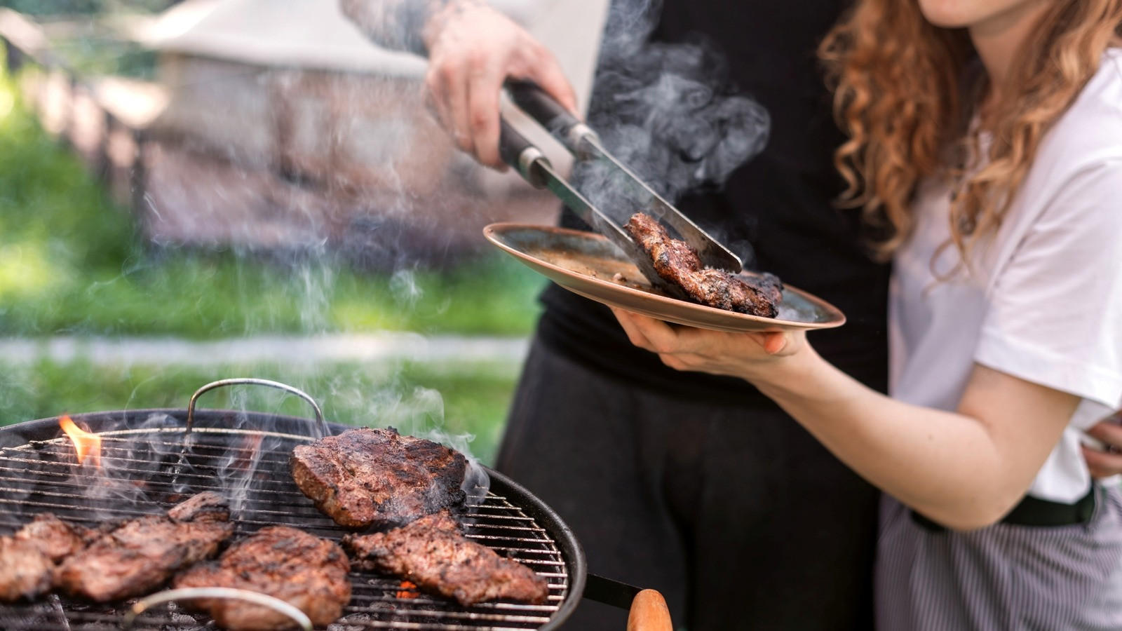 Two people serving grilled food together at an outdoor barbecue.
