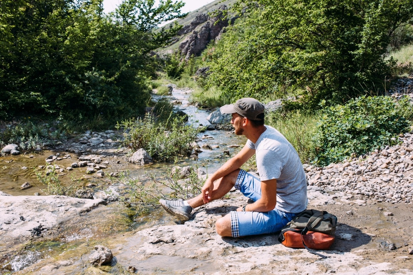 Man sitting beside a stream outdoors, taking a quiet pause in a natural setting.
