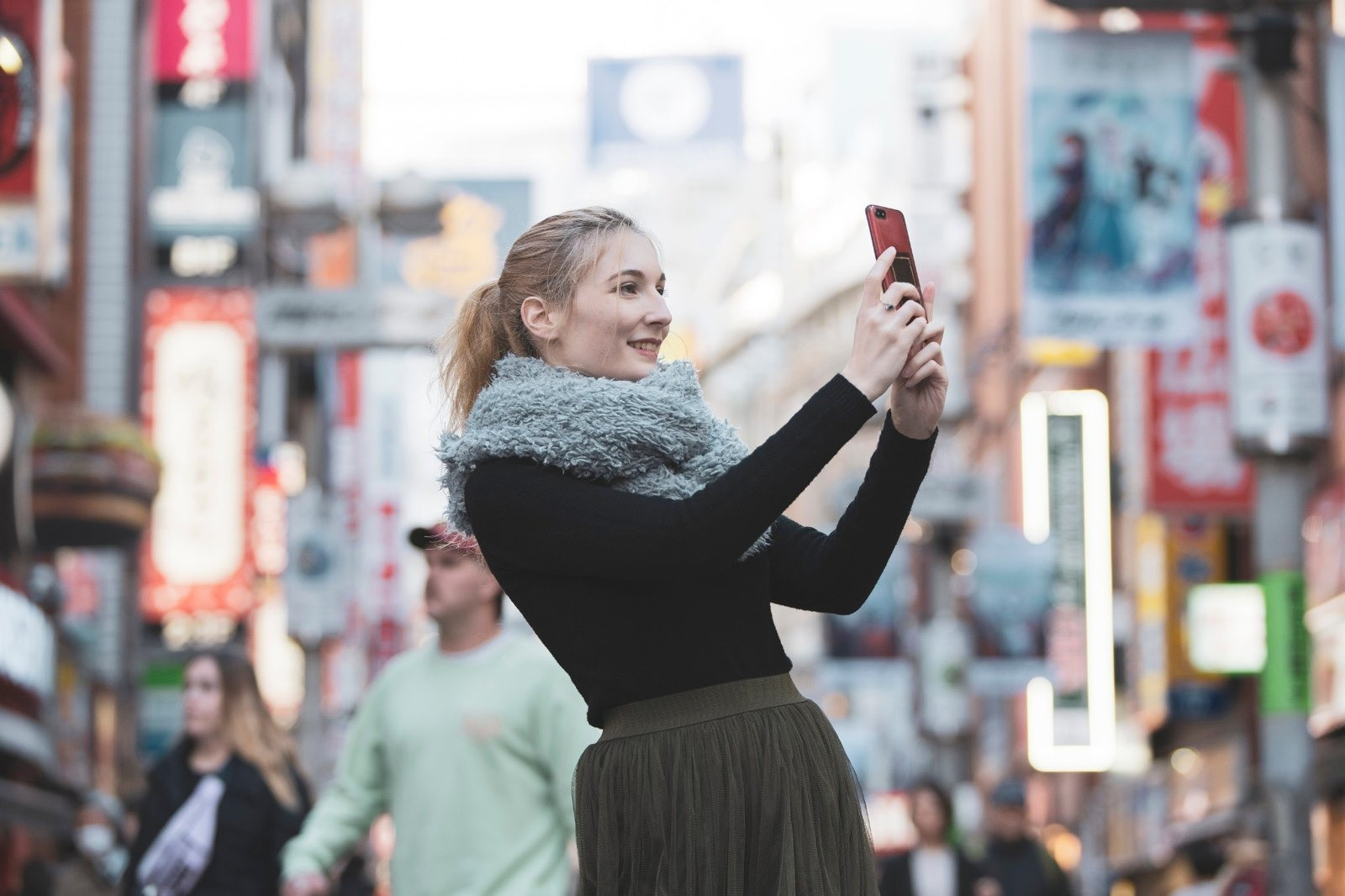 Woman wearing a gray scarf and holding up a smartphone on a busy city street.