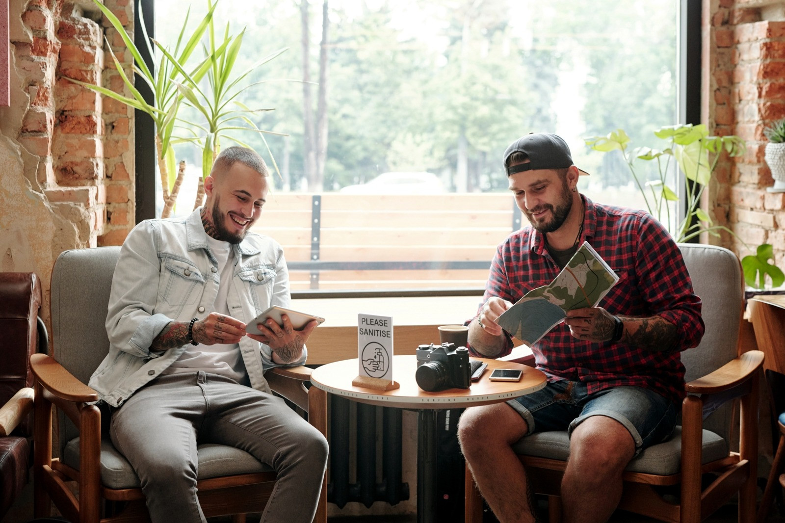 Two men seated beside a café window, one using a tablet and the other looking at a folded map.