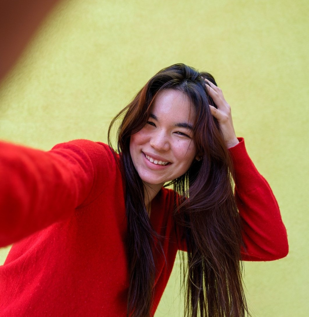 Smiling woman with long dark hair wearing a red sweater and taking a selfie against a yellow-green wall.