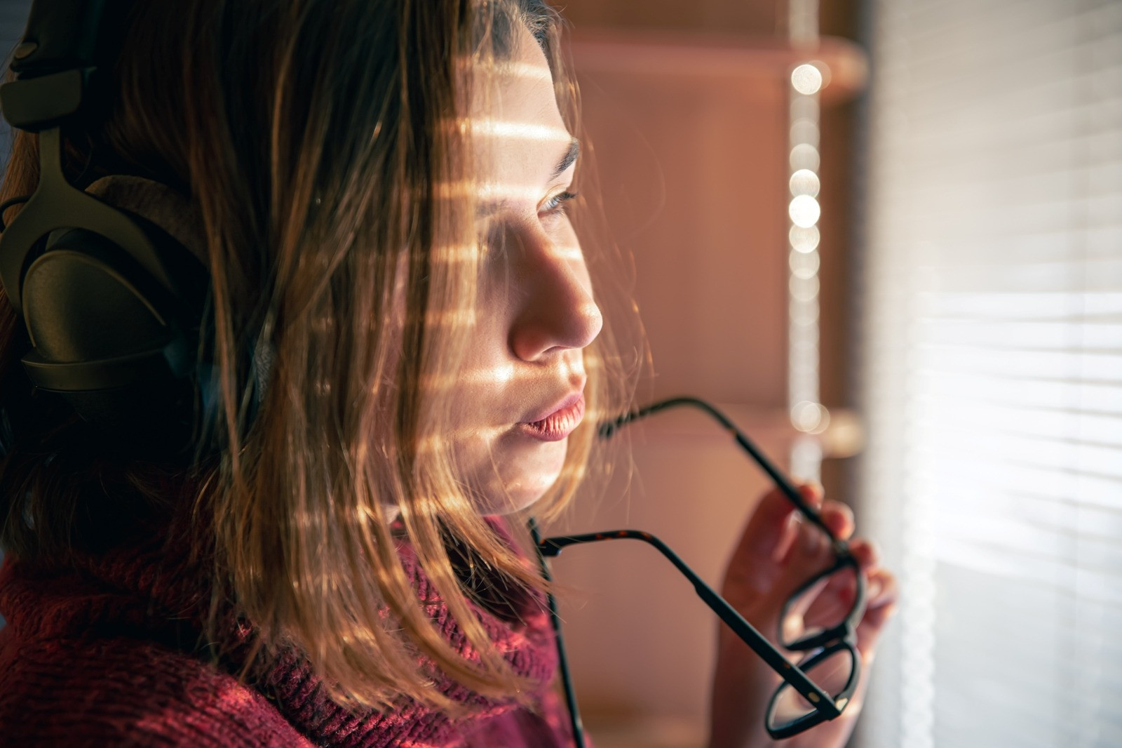 Close-up of a woman wearing headphones and holding eyeglasses while looking thoughtfully out a sunlit window, taking a quiet moment to herself.