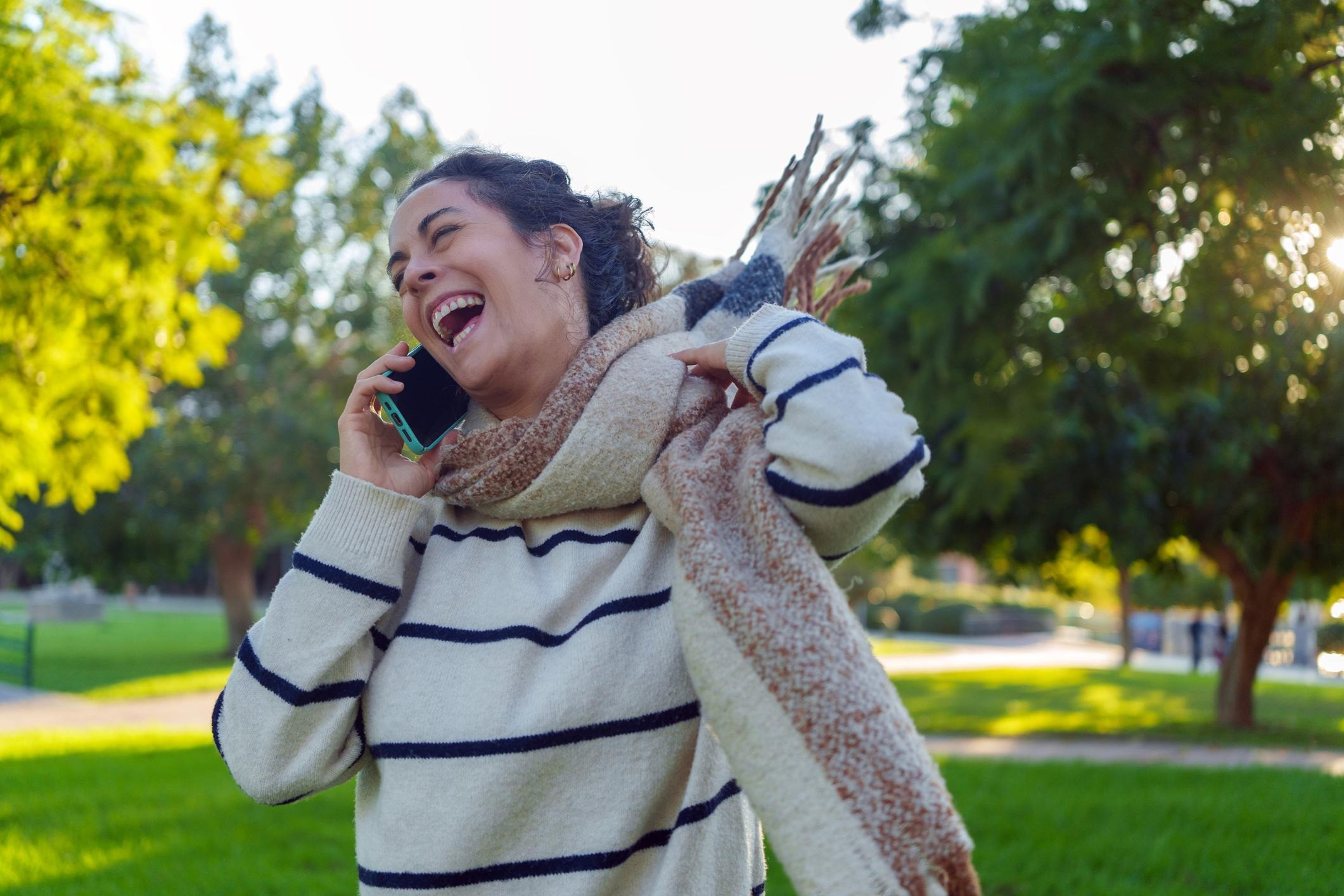 Woman laughing while talking on a smartphone outdoors, wearing a striped sweater and a large scarf.