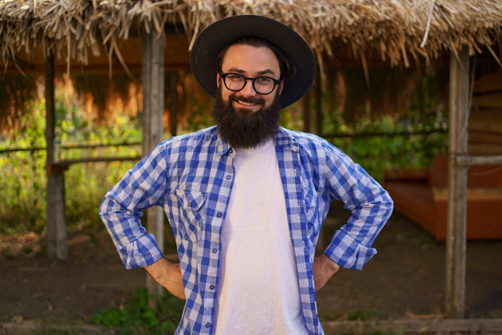 Smiling bearded man wearing glasses, a black hat, and a blue plaid shirt standing outdoors in front of a rustic building.