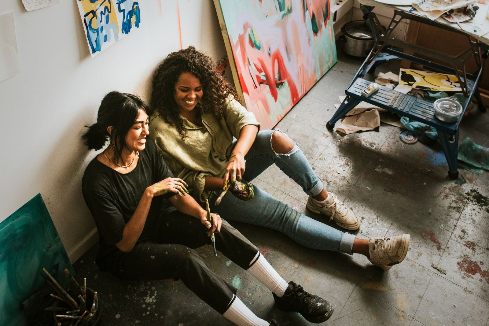 Two smiling women sitting together in a colorful art studio, holding paintbrushes and enjoying a relaxing creative moment.