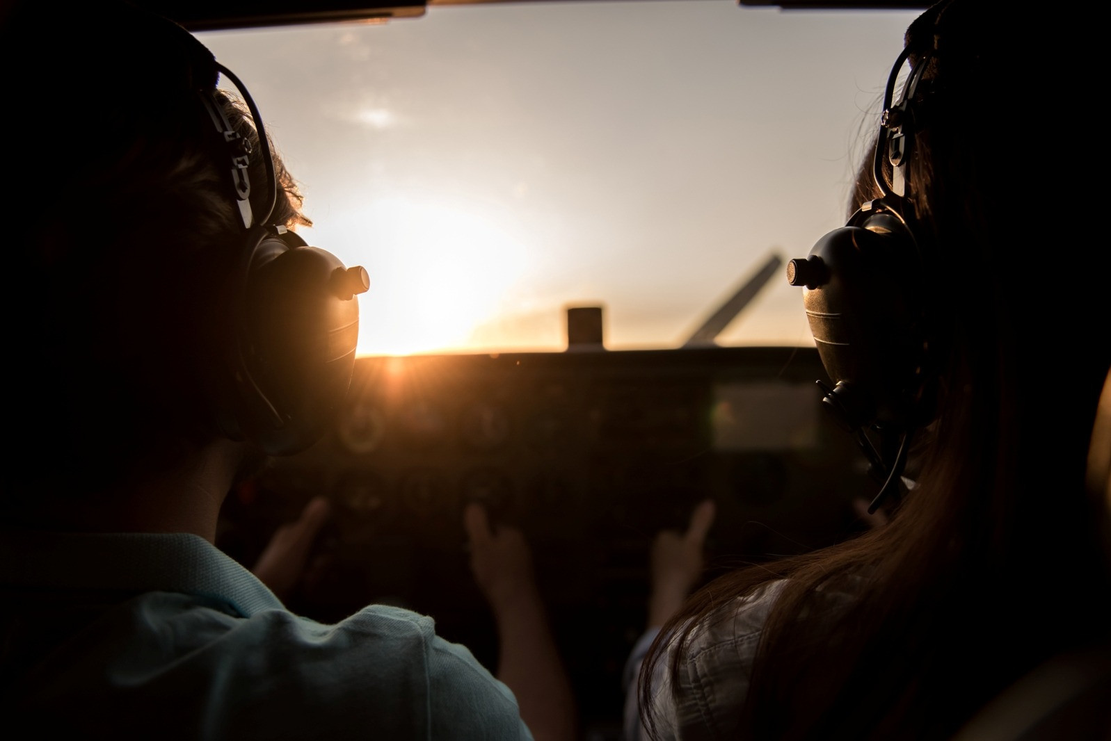 Two people wearing aviation headsets sit in an aircraft cockpit with sunlight shining through the windshield.