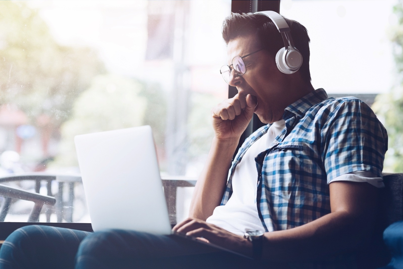 Man wearing headphones and looking at a laptop while seated indoors near a window.