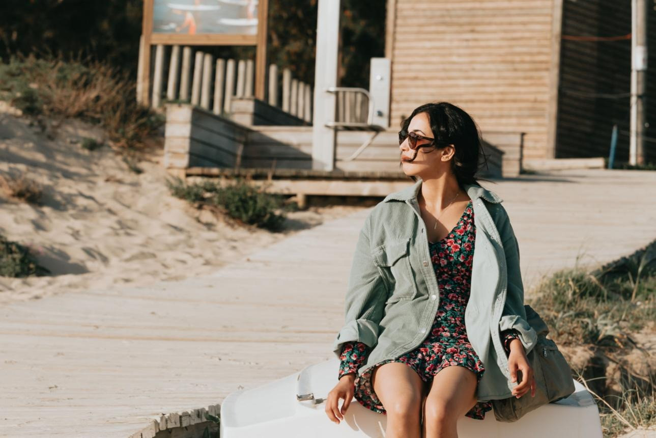 Woman wearing sunglasses and a green jacket sitting outdoors near a beachside building.