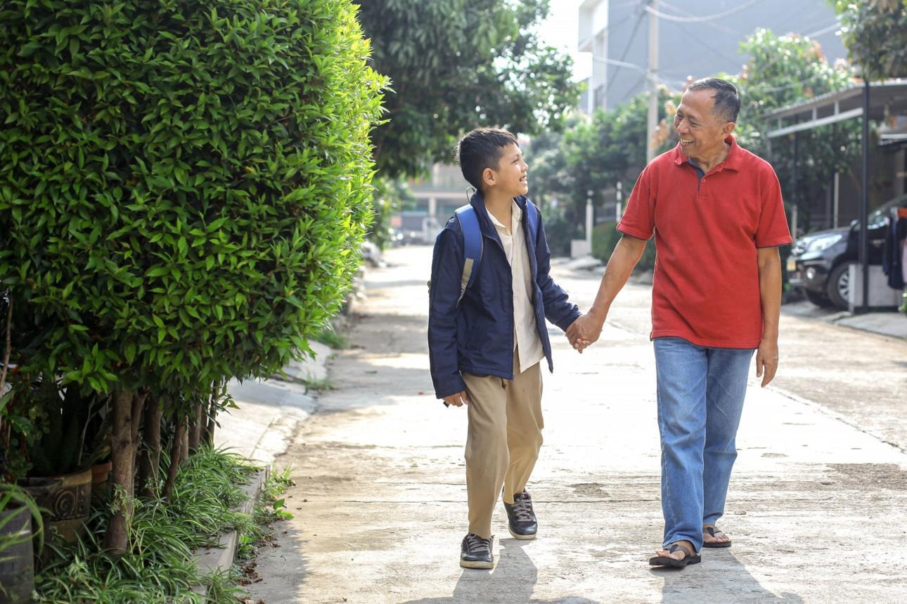 Older man in a red shirt walking hand in hand with a school-aged boy carrying a backpack along a sunny residential street.