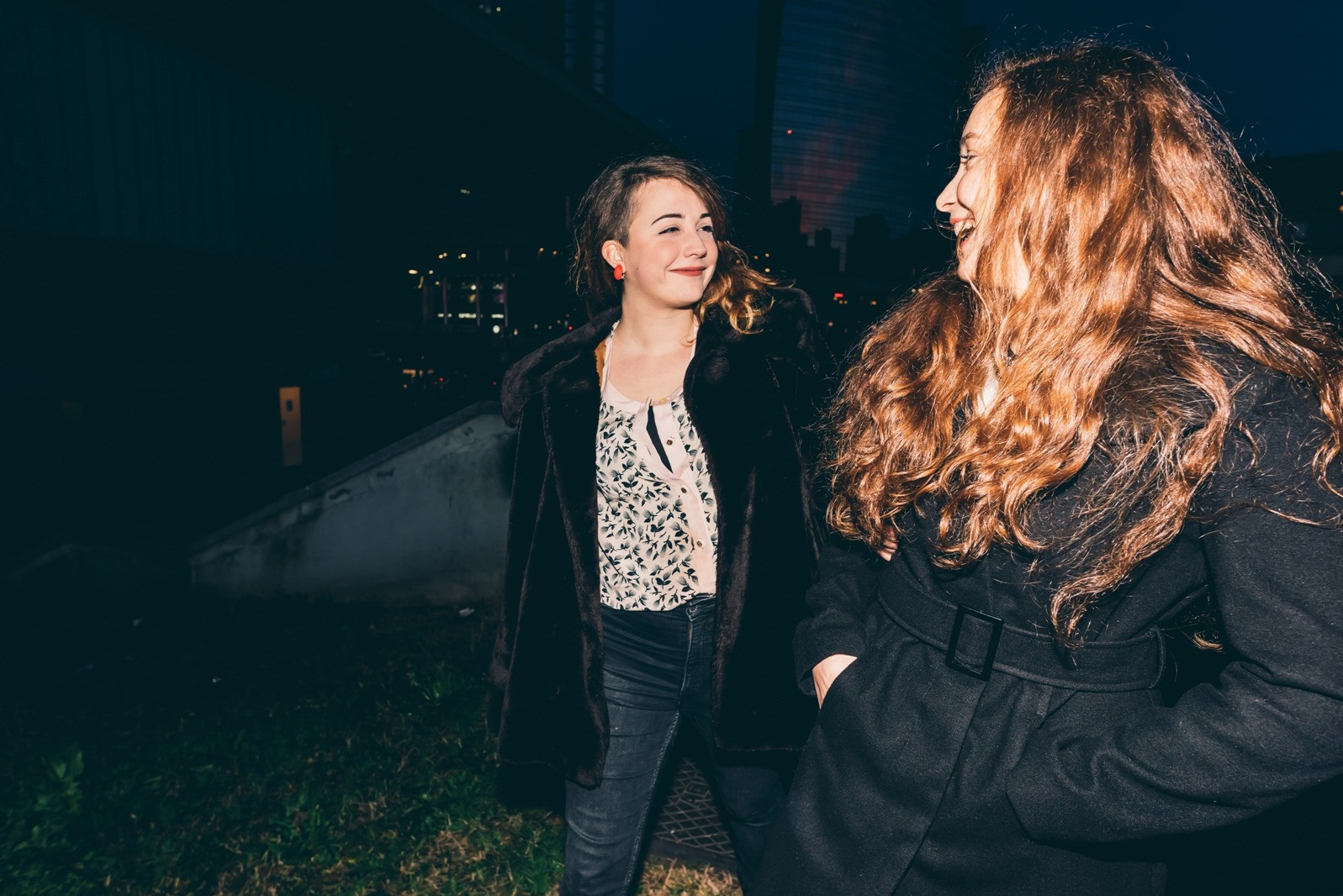 Two women wearing dark coats smile at each other while standing outdoors at night.